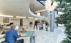 Öppen kontorsyta med modern inredning, en svängd trappa och stora fönster. En person sitter vid ett skrivbord och arbetar vid en dator och en annan person går. Olika växter och konstverk är synliga.