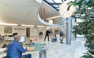 Modernt kontor med mezzanin och marmorgolv. En man sitter vid ett skrivbord med en dator, medan en annan man arbetar vid ett bord i bakgrunden. Växter och modern belysning syns.