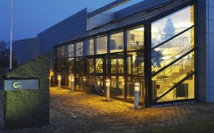 En modern byggnad med glasväggar i skymningen, med upplyst interiör som visar flera våningar och en trappa. En upplyst yttre skylt och gångvägsljus kantar kullerstensgången.