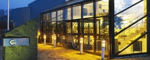 En modern glasbyggnad i skymningen med upplyst interiör, med stora fönster, en trappa och en skylt utanför.