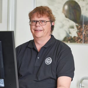 En person som bär glasögon och ett headset sitter vid ett skrivbord med en datorskärm framför sig och ler. De är klädda i en svart pikétröja.