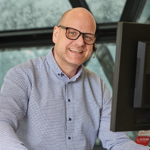 En man med glasögon och ett headset ler vid sitt skrivbord medan han arbetar på en dator.