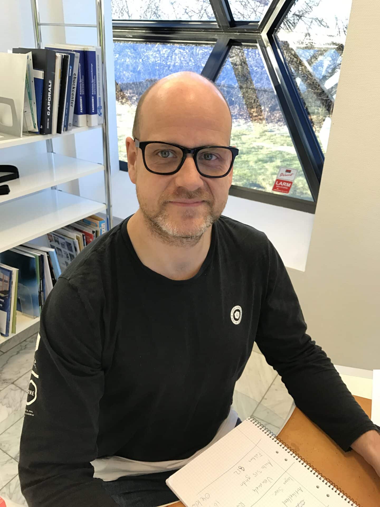 En person med glasögon och svart skjorta sitter vid ett skrivbord med en anteckningsbok, framför ett geometriskt fönster med bokhyllor i bakgrunden.