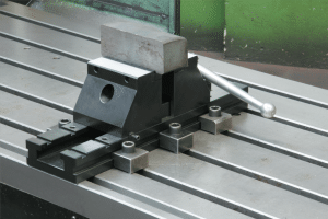 Ett metallskruvstycke med ett materialblock kläms fast på en randig metallyta, med ett långt handtag som sträcker sig från höger sida.