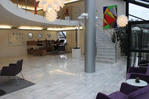 Modern kontorslobby med marmorgolv, en trappa och geometriska konstverk på väggarna. En receptionsdisk är bemannad av personal och moderna möbler är snyggt ordnade i sittgruppen.